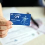 Close-up de uma mão segurando um cartão azul de CPF (Cadastro de Pessoas Físicas) em primeiro plano. Ao fundo, há documentos sobre uma mesa, notas de dinheiro desfocadas e outros objetos de escritório fora de foco. O cartão exibe o logotipo da Receita Federal e informações de exemplo, enquanto a profundidade de campo reduzida destaca o documento. A iluminação é suave e o ambiente sugere contexto administrativo ou financeiro.