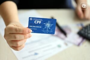 Close-up de uma mão segurando um cartão azul de CPF (Cadastro de Pessoas Físicas) em primeiro plano. Ao fundo, há documentos sobre uma mesa, notas de dinheiro desfocadas e outros objetos de escritório fora de foco. O cartão exibe o logotipo da Receita Federal e informações de exemplo, enquanto a profundidade de campo reduzida destaca o documento. A iluminação é suave e o ambiente sugere contexto administrativo ou financeiro.